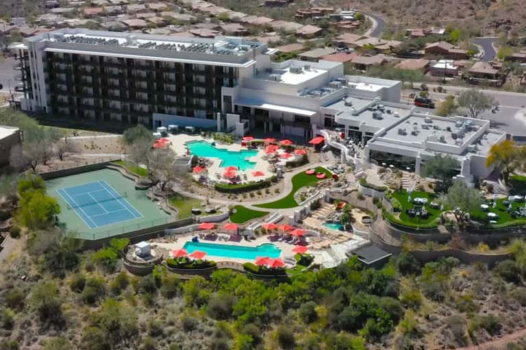 Aerial view of modern resort with turquoise pools, red umbrellas, tennis court, and surrounding desert landscape