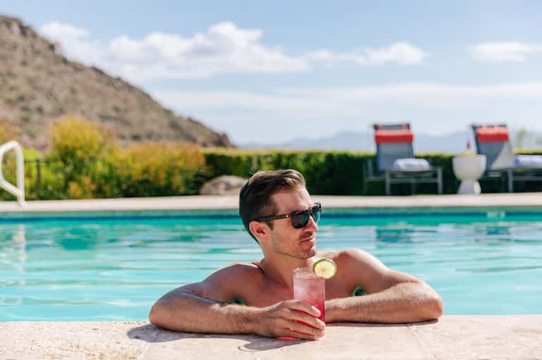 Man in sunglasses holding pink cocktail while floating in resort pool with desert backdrop