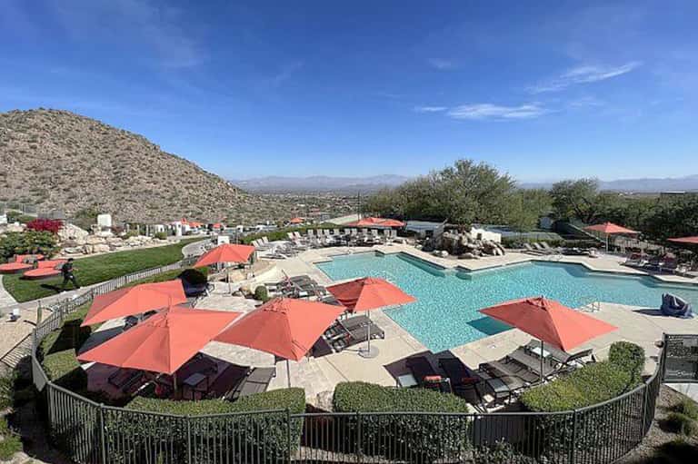 Desert resort pool complex with coral umbrellas, loungers, mountain views, and surrounding vegetation