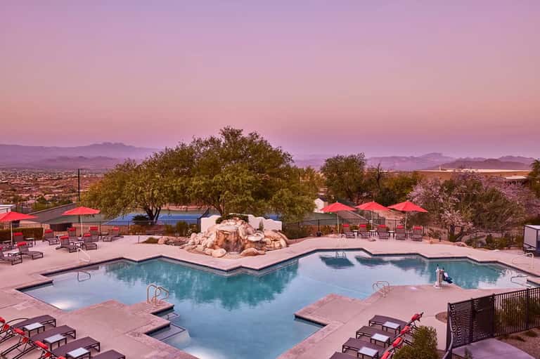 Resort pool at dusk with red umbrellas, lounge chairs, desert landscape, and pink sky