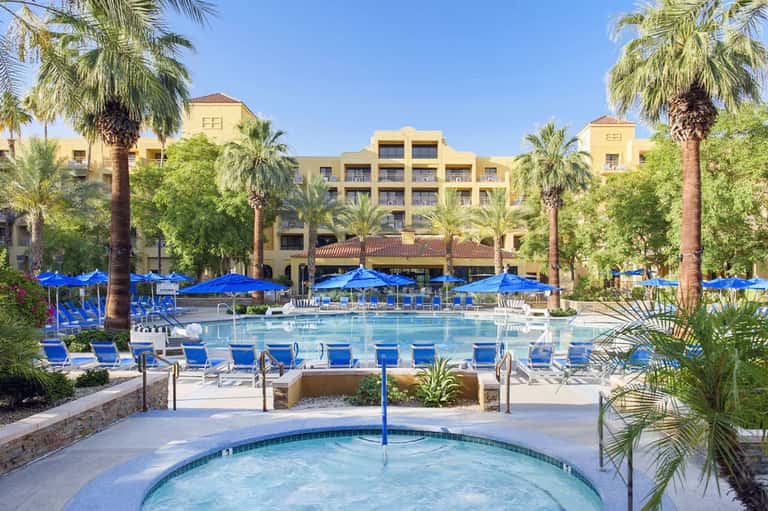 Resort pool complex with blue umbrellas, lounge chairs, palm trees, and yellow hotel building
