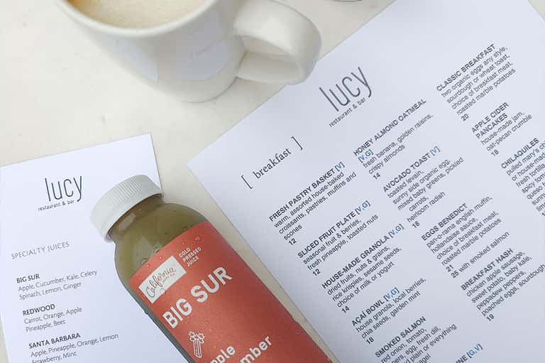 Restaurant menu and specialty juice bottle at cafe table with white mug