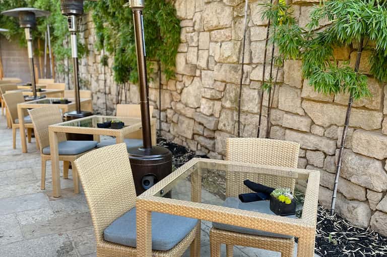 Outdoor patio with woven chairs and glass-top tables against stone wall with greenery