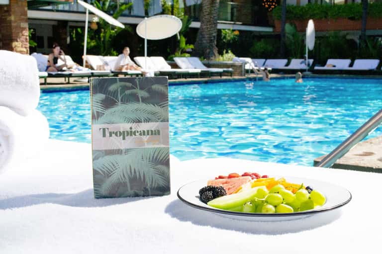 Poolside table with fresh fruit plate and Tropicana drink menu overlooking blue swimming pool