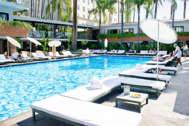 Resort pool with white lounge chairs, umbrellas, and palm trees surrounded by modern buildings