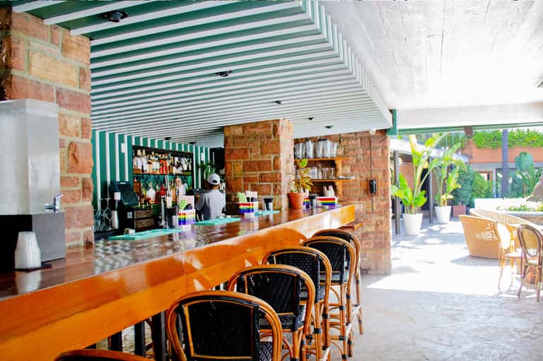 Outdoor resort bar with wooden counter, rattan chairs, and striped awning under brick archway