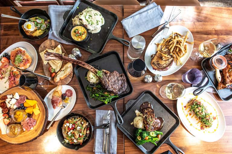 Overhead view of restaurant table with multiple dishes, steaks, sides, cocktails, and shared plates