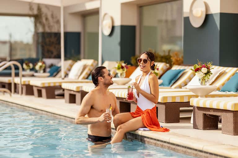Couple enjoying pool area with striped lounge chairs, water features, and modern resort architecture