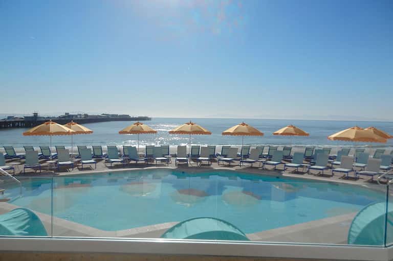 Beachfront pool with tan umbrellas, lounge chairs, glass barrier, and ocean pier backdrop on sunny day