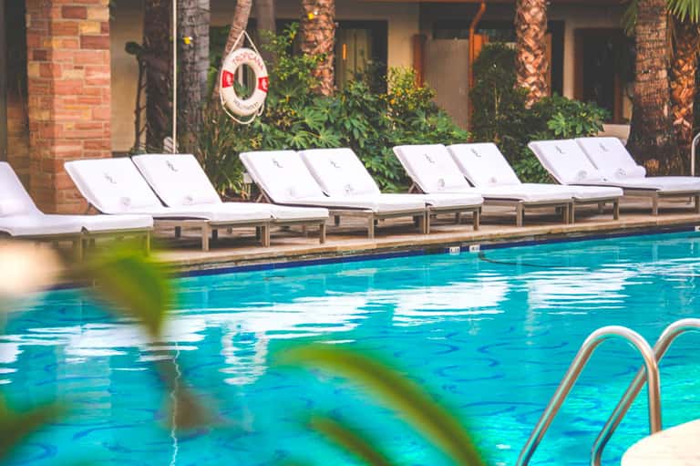 Resort pool with turquoise water, white loungers with cushions, palm trees, and brick building with ivy
