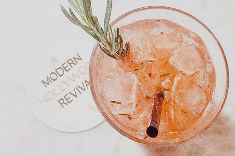 Peach-colored cocktail with rosemary garnish and Modern Holliwax Revival text