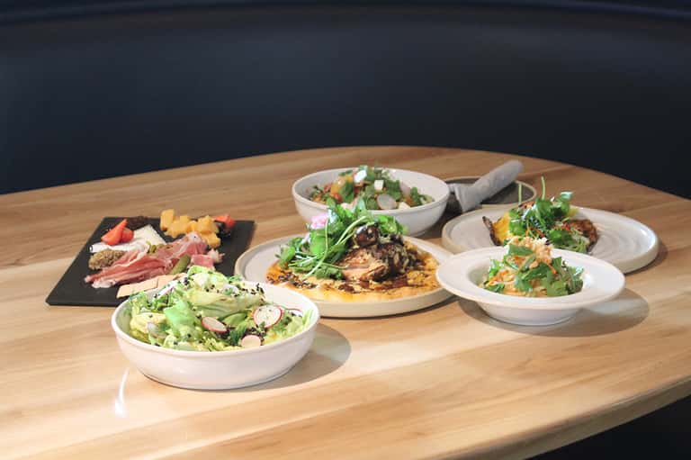 Gourmet plated dishes on wooden table including salads, cured meats, pizza, and appetizers with dark backdrop