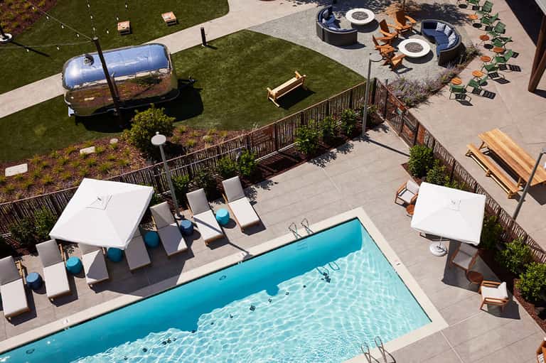Modern resort pool area with white umbrellas, loungers, Airstream trailer, and fire pits on lawn