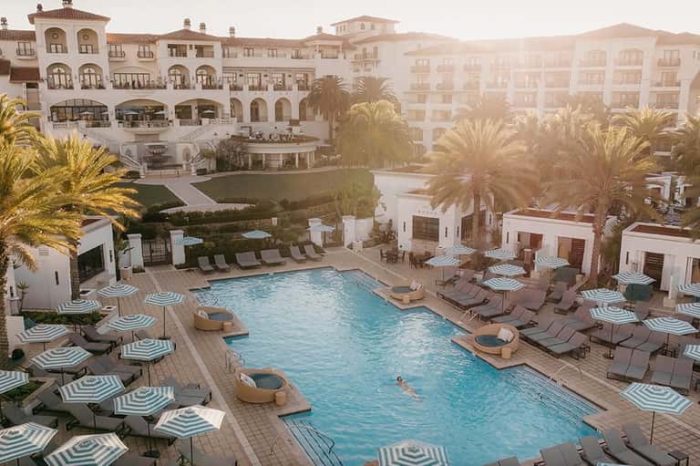 Mediterranean-style resort pool with striped umbrellas, loungers, archways, and palm-lined grounds