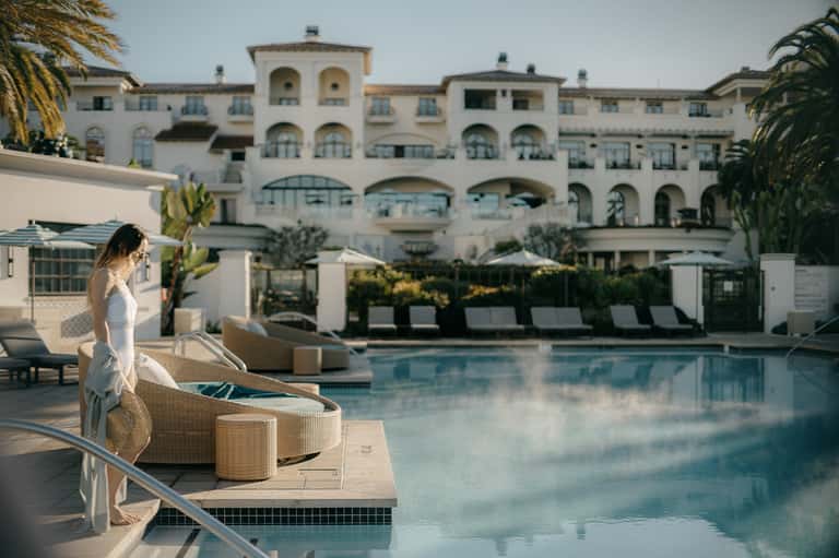 Woman in white dress at luxury resort pool with Mediterranean architecture and palm trees in background