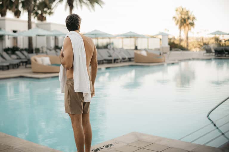 Man standing poolside at resort with palm trees and lounge chairs in background