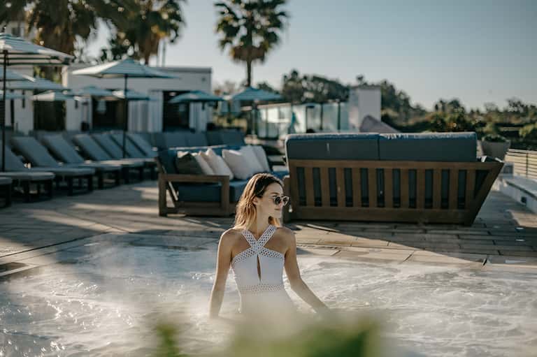 Woman in white swimsuit enjoying outdoor heated pool with resort buildings and palm trees visible