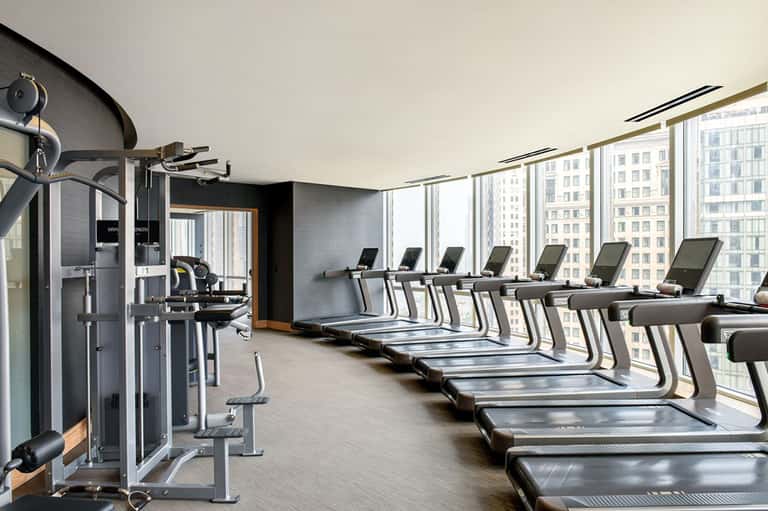 High-rise gym with rows of treadmills facing floor-to-ceiling windows overlooking city skyline