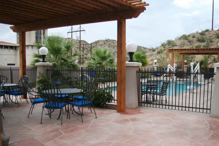 Covered patio seating area with wooden pergola, metal chairs, and pool view beyond fence