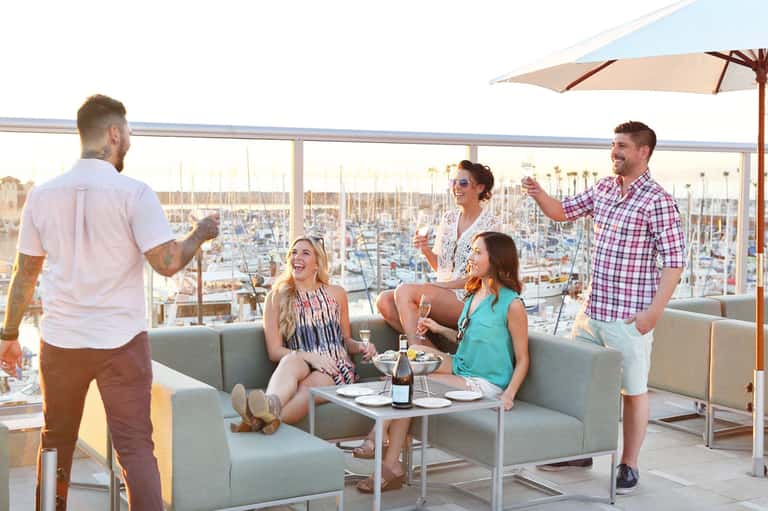Group of friends enjoying drinks on a rooftop terrace overlooking a marina with sailboats