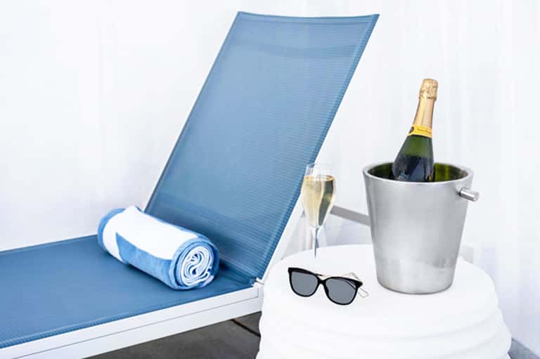 Lounge chair with blue cushion, champagne bucket, sunglasses, and rolled towel on white table