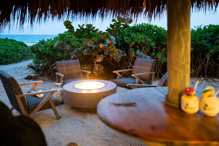Beach fire pit lounge with woven chairs under thatched roof at sunset with ocean beyond