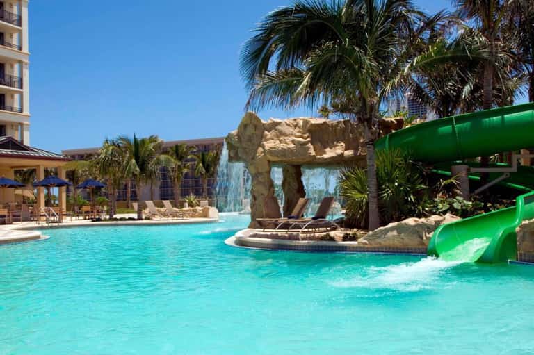 Resort pool with turquoise water, rock archway feature, green waterslide, and tropical landscaping