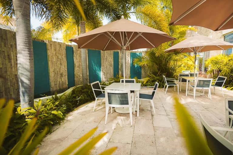 Shaded outdoor patio with white umbrellas, palm trees, and dining tables in a resort setting
