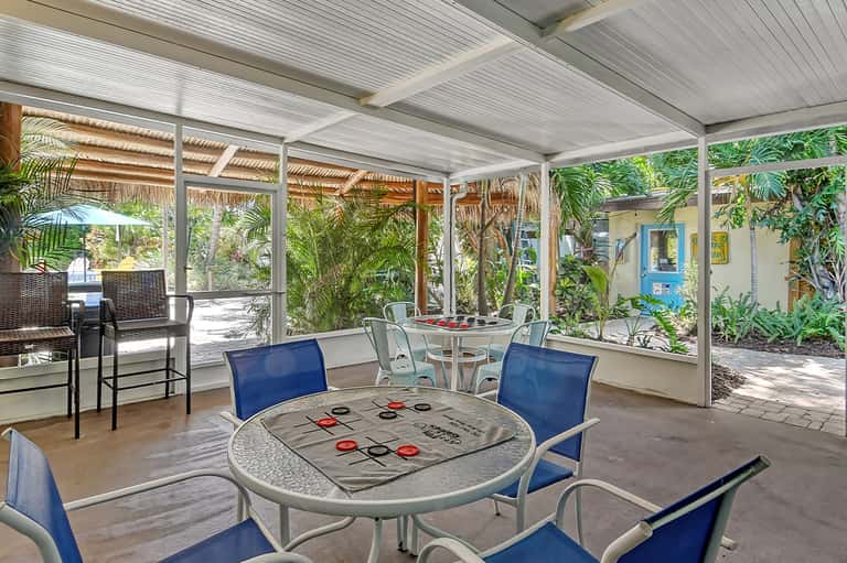 Covered patio lounge area with tic-tac-toe table, chairs, and tropical vegetation views