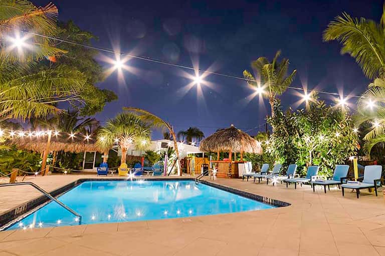 Illuminated resort pool at night surrounded by palm trees, thatched structures, and lounge chairs