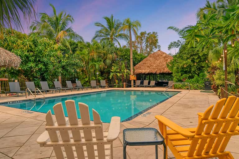 Resort pool surrounded by Adirondack chairs, thatched structures, and tropical vegetation