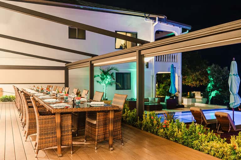 Modern residential deck at night with dining table, woven chairs, and glowing pool view