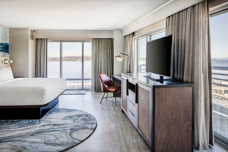 Waterfront hotel bedroom with king bed, TV, burgundy chair, and panoramic balcony water views