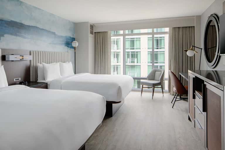 Contemporary hotel bedroom with two queen beds, blue accent wall art, floor-to-ceiling windows, and modern furnishings