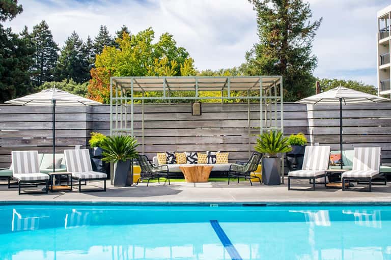 Resort pool deck with lounge chairs, umbrellas, and wooden privacy fence backdrop
