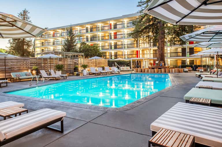 Illuminated hotel pool at dusk with striped loungers, wooden fencing, and multi-story building backdrop