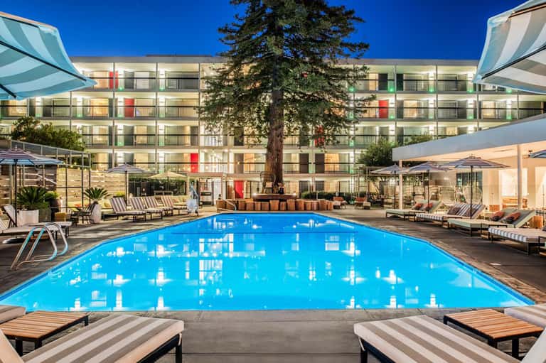 Resort pool at dusk with bright blue water, lounge chairs, umbrellas, and illuminated hotel building