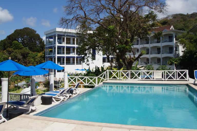 Resort pool with blue umbrellas, white colonial buildings, lounge chairs, and surrounding mature trees