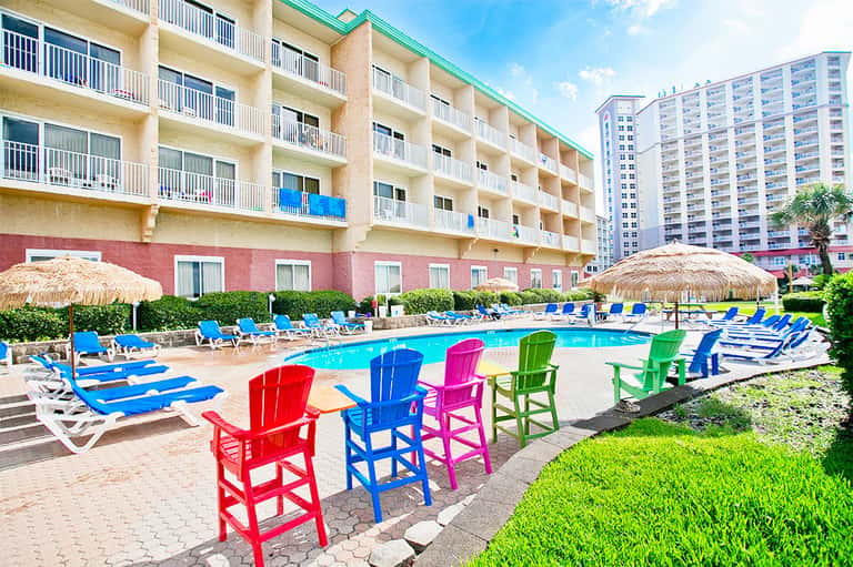 Resort pool area with colorful Adirondack chairs, thatched umbrellas, and multi-story beachfront hotel building
