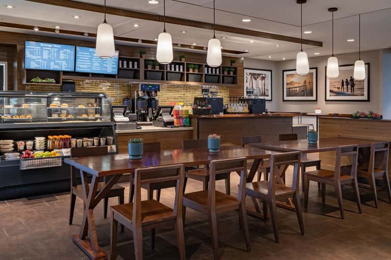 Contemporary hotel breakfast area with wooden tables, pendant lighting, bakery case, and menu board