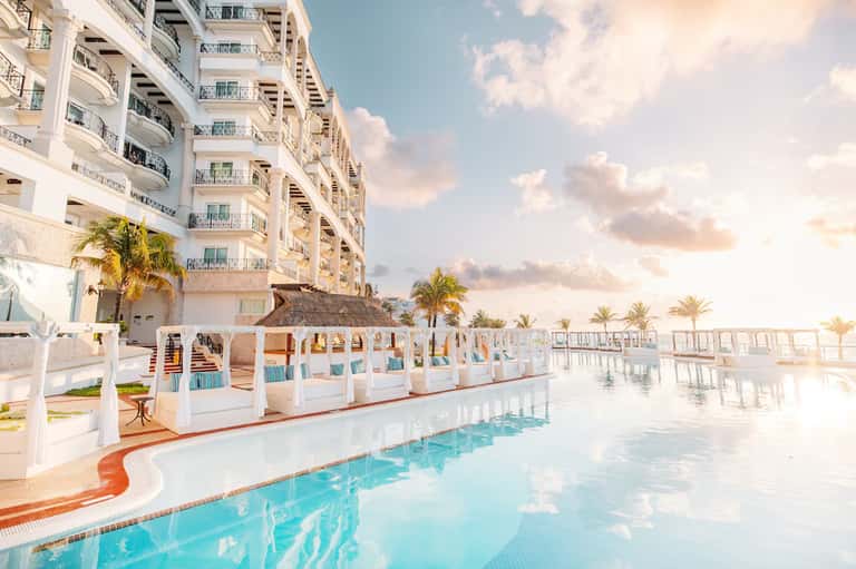 Luxury beachfront resort pool with cabanas, high-rise building, and sunset sky