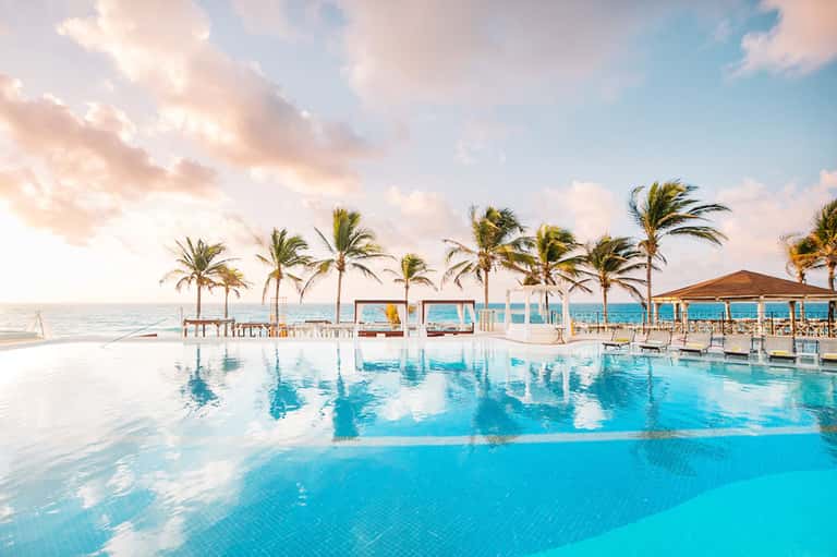 Oceanfront infinity pool with palm trees, white cabanas, and reflections under soft cloudy sky