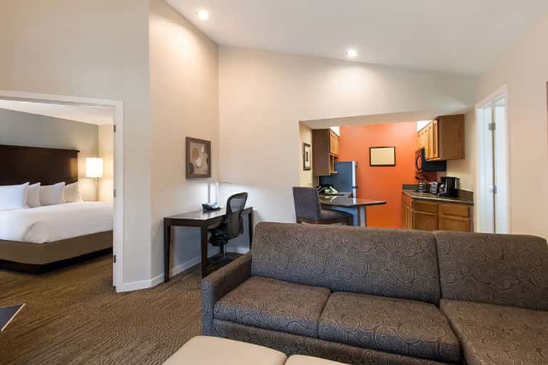 Modern hotel suite with bedroom, living area, kitchenette, desk, and orange accent wall