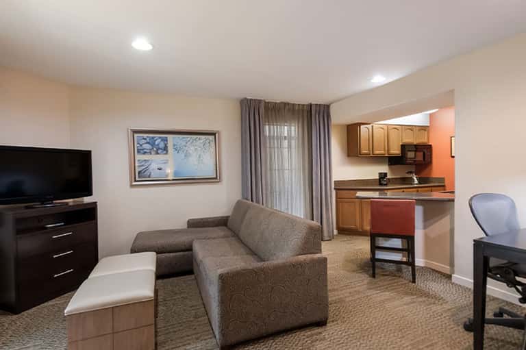 Hotel suite with sofa, kitchenette, TV, desk area, and warm neutral décor