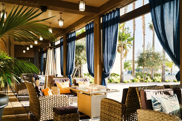 Covered patio seating area with woven furniture, blue curtains, wooden beams, and palm tree views