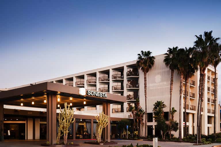 Modern beige hotel exterior with palm trees, illuminated entrance canopy, and 'SONESTA' sign at dusk