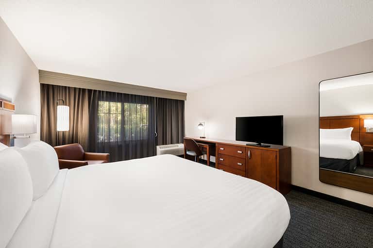 Modern hotel room with white bedding, wooden furniture, dark curtains, and flat-screen TV
