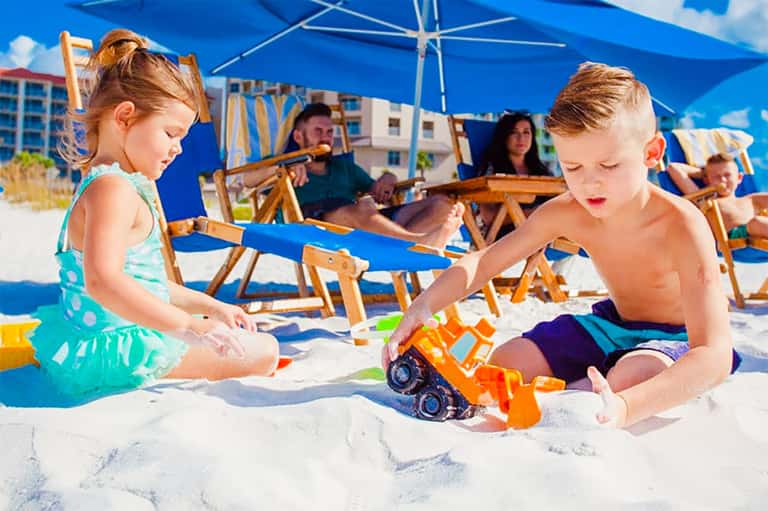 Young children playing with toy trucks in sand under blue beach umbrellas with parents nearby