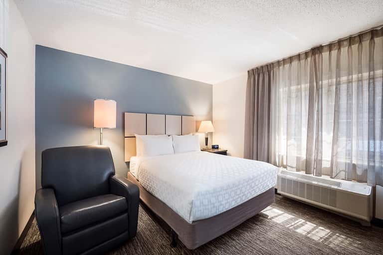 Contemporary hotel bedroom with queen bed, blue accent wall, dark leather chair, and floor-to-ceiling windows