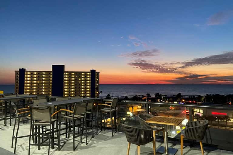 Rooftop bar with high-top seating overlooking illuminated beachfront resort and sunset ocean view